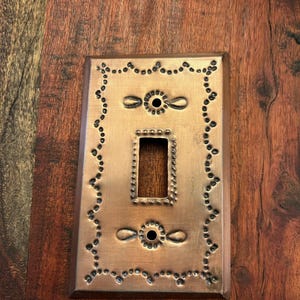 May include: Copper-colored light switch cover with a decorative design. The rectangular cover features a central opening for the switch and is adorned with a dotted border and decorative accents. The cover is made of metal and has a rustic appearance.