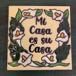 Puede incluir: Azulejo cerámico cuadrado con fondo crema. El azulejo presenta una corona de calas blancas con hojas verdes. El texto "Mi Casa es su Casa" está escrito en negro en el centro del azulejo.