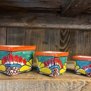 May include: Three colorful ceramic planters with a square shape. The planters have a vibrant design with red, yellow, and blue floral patterns on a green background, and orange rims. They are arranged on a wooden shelf.