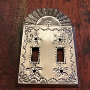 May include: A silver-colored, metal light switch cover with a decorative floral design. The cover has two switch openings and is adorned with embossed flowers and a sunburst-like top. The background is a dark wood surface.