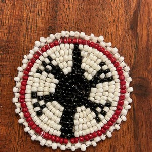 May include: A round beaded design with a black center surrounded by white beads and a red outer ring.