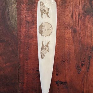 Puede incluir: Una talla de madera alargada de color crema con un extremo puntiagudo. Presenta imágenes grabadas de calaveras de animales y un diseño circular con texto. La talla se muestra sobre un fondo de madera oscura.