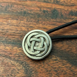 May include: A circular silver-toned metal hair tie accessory featuring an embossed, intricate, four-fold interlaced knot pattern. The metallic disc is securely attached to a thin, black elastic hair band, creating a functional hair accessory.