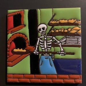 Puede incluir: Un azulejo de cerámica colorido que representa a un panadero esqueleto con un delantal azul, usando una pala de madera para hornear pan en un horno de ladrillo. El azulejo tiene un fondo verde y presenta un estante con panes.