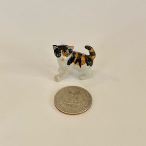May include: A small, ceramic calico cat figurine with black, orange, and white markings. The cat is in a walking pose, with a quarter coin next to it for size comparison. The figurine is detailed and appears to be hand-painted.
