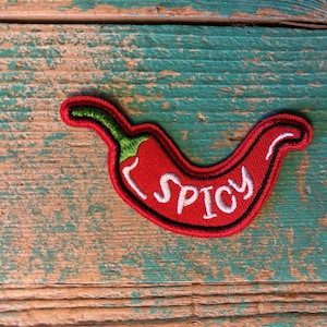 May include: A red chili pepper patch with the word "SPICY" embroidered in white. The patch has a green stem and a black border. The patch is on a weathered, turquoise-colored wooden surface.