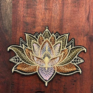 May include: Embroidered lotus flower patch with a detailed design. The patch features shades of gold, brown, and lavender, outlined in black. The patch is likely for clothing or accessories.