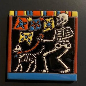 May include: A colourful ceramic tile featuring a skeleton holding a lead and walking a skeletal dog. The tile has a black background with a border of red, orange, yellow, blue and green. The skeleton is wearing a white shirt and has a black hat. The dog is wearing a red collar. The tile is decorated with colourful flags.