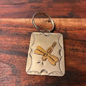 May include: Silver-tone metal keychain with a rectangular pendant. The pendant features a gold-colored dragonfly design in the center. The edges of the pendant have a decorative, scalloped pattern. The keychain ring is silver.