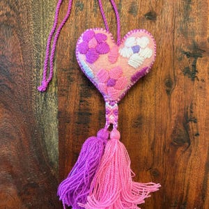 May include: A pink felt heart-shaped ornament with embroidered flowers in shades of pink and purple. It has a braided cord for hanging and two tassels in pink and purple. The ornament is displayed on a wooden surface.