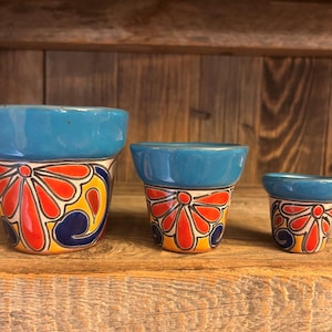 May include: Three ceramic flower pots in descending sizes, each featuring a solid blue rim and a hand-painted base with a vibrant floral pattern in red, yellow, and dark blue. The pots have a tapered shape and a smooth, glossy finish.