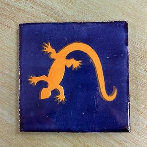 May include: A square tile with a blue background and an orange lizard silhouette.