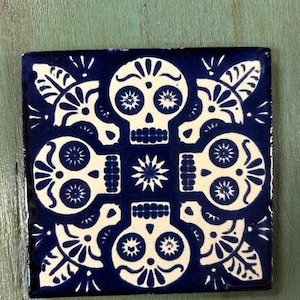 May include: A blue and white ceramic tile with a repeating pattern of four sugar skulls. The skulls are stylized with intricate details and have white eyes and a black outline.