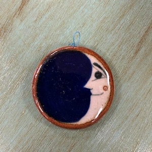 May include: A ceramic ornament shaped like a crescent moon with a stylized face. The moon is painted in shades of blue and white with a brown rim.