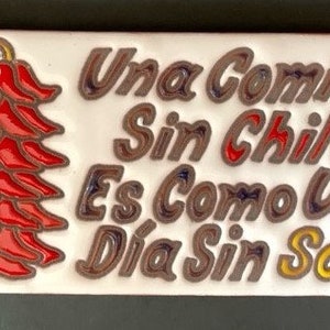 May include: Rectangular ceramic tile with a red chili pepper design on the left and Spanish text on the right. The text reads "Una Comida Sin Chile Es Como Un Día Sin Sol."