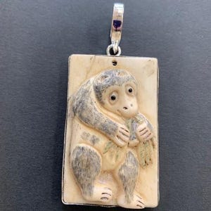 Op de afbeelding: Een rechthoekige hanger met een zilveren beugel met een gebeeldhouwde aap die een bladrijke tak vasthoudt. De aap is afgebeeld in grijstinten en crème, tegen een crèmekleurige achtergrond. De hanger is omlijst met een zilveren rand.