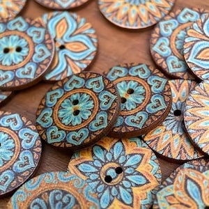 Op de afbeelding: Houten knopen met een bloemmotief in tinten van teal, oranje en bruin. Elke knoop heeft twee gaten om op te naaien. De knopen zijn gerangschikt op een houten oppervlak. Diameter ongeveer 3 cm.