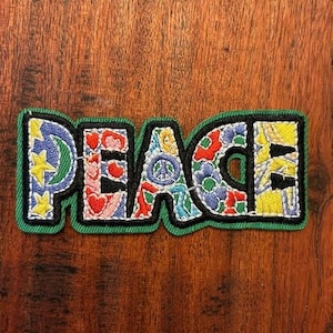 May include: Embroidered patch with the word "PEACE" in colorful, floral-patterned letters. The patch has a green border and black outline, with each letter featuring a unique design, including stars, hearts, and a peace symbol.