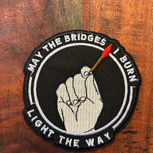 May include: Black and white embroidered patch with a white hand holding a red and yellow match. The patch has a white circle with the text "May the bridges I burn light the way" around the edge.