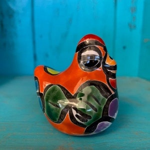 May include: A small, hand-painted ceramic chicken figurine with a bright orange body, black and white eyes, and green, purple, and yellow accents.