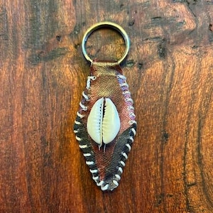 Puede incluir: Un llavero de cuero marrón con una concha de cauri blanca en el centro. El llavero tiene un borde cosido blanco y un anillo de metal.