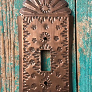 May include: A copper-colored metal light switch cover with a decorative floral design. The cover has a rectangular shape with a single switch opening and a fan-shaped top. The background is weathered turquoise wood.