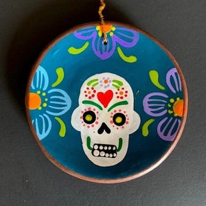 May include: A teal blue ceramic ornament with a painted sugar skull design. The skull has a red heart, yellow eyes, and white teeth. The ornament is decorated with colorful flowers and has a gold string for hanging.