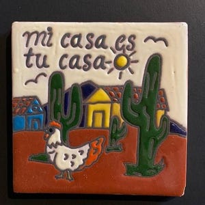 May include: A ceramic tile with a colorful illustration of a house, cacti, a chicken, and the text "mi casa es tu casa" in Spanish. The tile has a white background with a red, orange, yellow, green, blue, and black color scheme.
