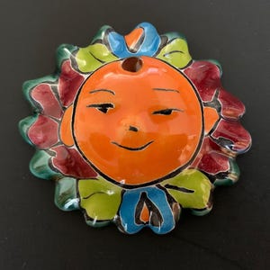 Könnte beinhalten: Ein keramisches, sonnenförmiges Dekorationsobjekt mit einem lächelnden Gesicht. Die Sonne hat ein orangefarbenes Gesicht, umgeben von bunten Blütenblättern in Rot, Grün und Blau. Das Stück ist wahrscheinlich handgefertigt und hat eine glänzende Oberfläche, ideal für die Inneneinrichtung.