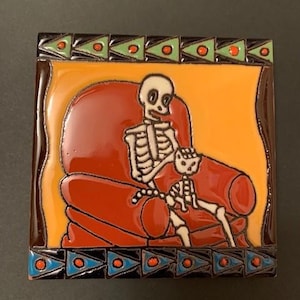 Op de afbeelding: Een keramische tegel met een kleurrijke rand en een skelet dat in een rode fauteuil zit met een kleine skeletkat. De achtergrond is oranje.