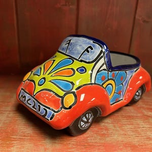 以下が含まれることがあります： 鮮やかな色のセラミック製の車型プランター。車は赤、黄、青、オレンジなどの明るい色で塗装されています。車の前面には「MCLII」の文字と屋根に数字の「1」があります。車輪は黒で、内装は暗いです。