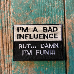 May include: A rectangular patch with the text "I'M A BAD INFLUENCE" above "BUT... DAMN I'M FUN!!!" in white lettering. The top section is off-white, and the bottom is black. The patch is on a weathered, turquoise wooden surface.