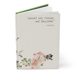 Peut inclure: Un journal avec une couverture beige clair portant la citation "WHAT WE THINK, WE BECOME" de Bouddha. La couverture présente également un motif floral à l'aquarelle avec des fleurs roses et blanches, des feuilles vertes et un papillon. Le journal a un dos vert.