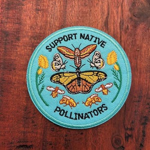 Könnte beinhalten: Ein runder, türkisfarbener Aufnäher mit der Aufschrift "SUPPORT NATIVE POLLINATORS". Das Design zeigt Schmetterlinge und Bienen in Orange, Gelb und Weiß, mit floralen Akzenten.