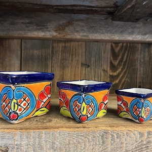 May include: Three square ceramic planters with a vibrant, hand-painted design. The planters feature a blue rim, orange background, and colorful floral patterns in red, yellow, and blue. They are arranged in a row, decreasing in size from left to right.
