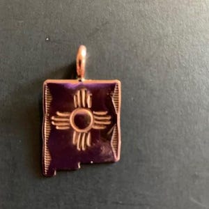 Peut inclure: Un pendentif en forme d'État du Nouveau-Mexique de couleur dorée avec le symbole de l'État, le symbole du soleil Zia, au centre.