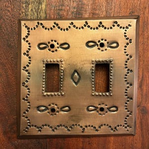 May include: A copper-colored, rectangular light switch plate with two switch openings. The plate features decorative punched-hole and embossed designs, including a diamond shape and floral-like accents. The edges are framed with a darker border.