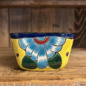 Puede incluir: Un pequeño plato de cerámica amarillo con un diseño floral azul y verde. El plato tiene un borde azul oscuro y está decorado con un centro rojo y blanco.