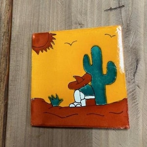 May include: A hand-painted ceramic tile featuring a yellow background, a red sun, a green cactus, and a cartoon figure wearing a red hat, sitting beneath the cactus.