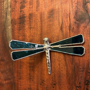 May include: A teal blue stained glass dragonfly with silver metal accents. The dragonfly is mounted on a wooden surface.
