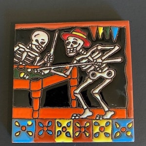 May include: A colorful ceramic tile with a Day of the Dead theme. Two skeletons are playing pool on a green felt table. The tile has a black background with orange, yellow, blue, and red accents.
