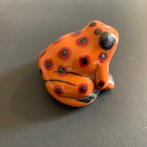 Könnte beinhalten: Orangefarbene Keramik-Froschfigur mit schwarzen und roten Flecken. Der Frosch sitzt auf seinen Hinterbeinen, die Vorderbeine sind eingezogen.
