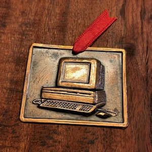 May include: A bronze-colored computer ornament with a red ribbon for hanging. The ornament features a detailed depiction of an old-style computer with a monitor, keyboard, and mouse. The ornament is rectangular with a textured surface.