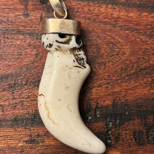 May include: A pendant featuring a curved, off-white tooth-shaped element carved into the likeness of a skull at the top. The piece is capped with a smooth, metallic silver-toned bail at the crown, designed for attachment to a necklace chain.