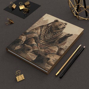 May include: A hardbound journal with a sepia-toned illustration of a bear in Native American attire, surrounded by bear cubs. The journal has a brown cover and is accompanied by gold and black pencils and paper clips.