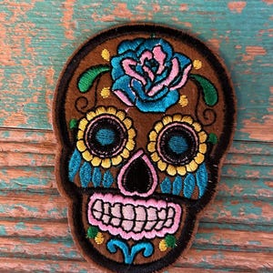 May include: Embroidered brown felt sugar skull patch with a blue and pink rose, yellow and blue eyes, and a pink and green mouth.
