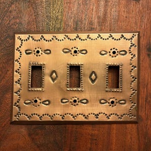 May include: A copper-colored light switch plate with three rectangular openings and two diamond-shaped accents. The plate has a decorative border with small, evenly spaced dots and floral-like designs. The plate is made of metal and has a rustic appearance.