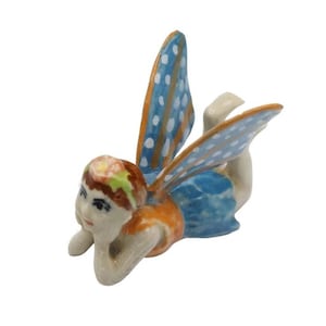 May include: A ceramic fairy figurine with blue and orange wings, a blue skirt, and an orange top. The fairy is lying down, with its head resting on its arm. The wings have white polka dots. The fairy's hair is a mix of colors.