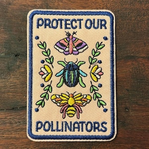 May include: A rectangular embroidered patch with a light beige background and a dark blue border. It features the text PROTECT OUR POLLINATORS in blue, with a moth, a beetle, and a bee centered between green leafy branches and small flowers.