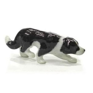 Puede incluir: Una figurita de porcelana negra y blanca de un perro Border Collie en pose de caza. El perro está de pie sobre sus cuatro patas con la cabeza baja y las orejas levantadas.